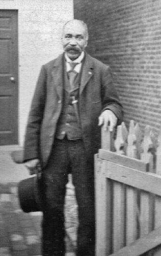 Photo of Benjamin Franklin Jennings standing outside a home, holding a fence with his hat in the other hand